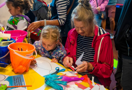 A photo of participants at a Coro Craft Morning. A young girl is colouring in a fish while an older female relative sits next to her working with some paper shapes.