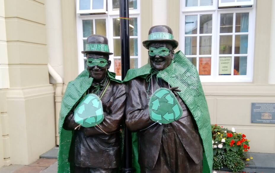 A photo of the Laurel and Hardy statue outside The Coro decked out as eco warriors, with green tarpaulin capes, small green shields with the universal symbol for recycling, green masks and green ribbons on their bowler hats