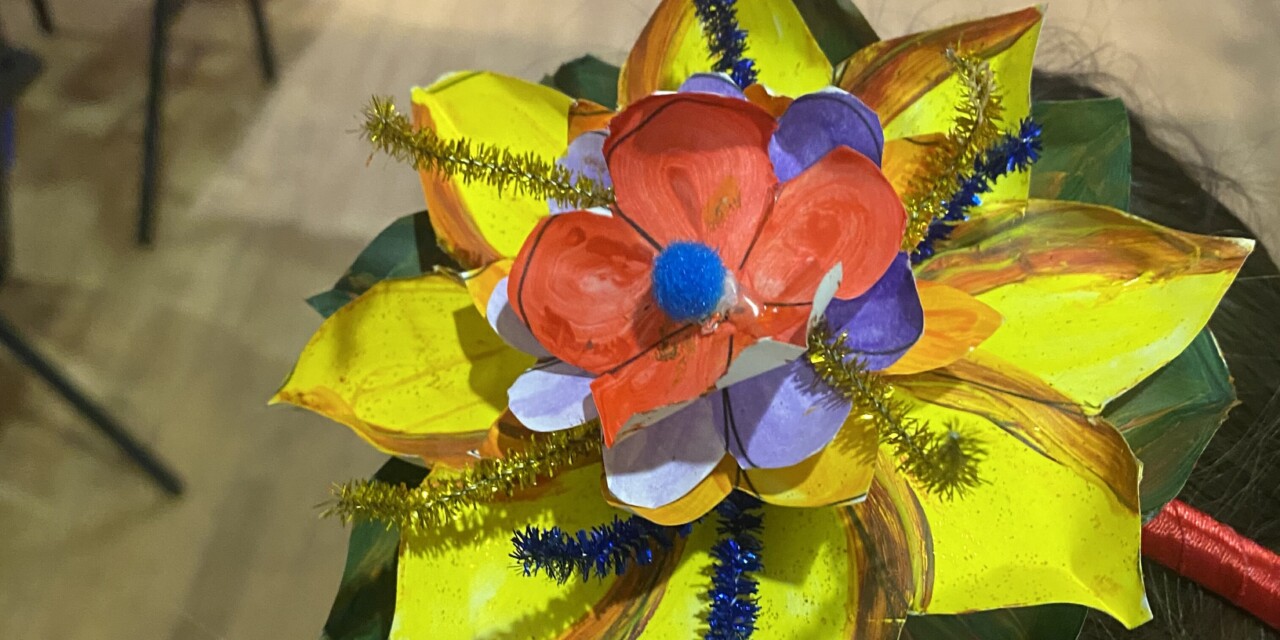 A photo of a mutli-coloured, hand-crafted headdress in the shape of a multi-tiered flower attached to a headband, made from coloured paper and tinsel.