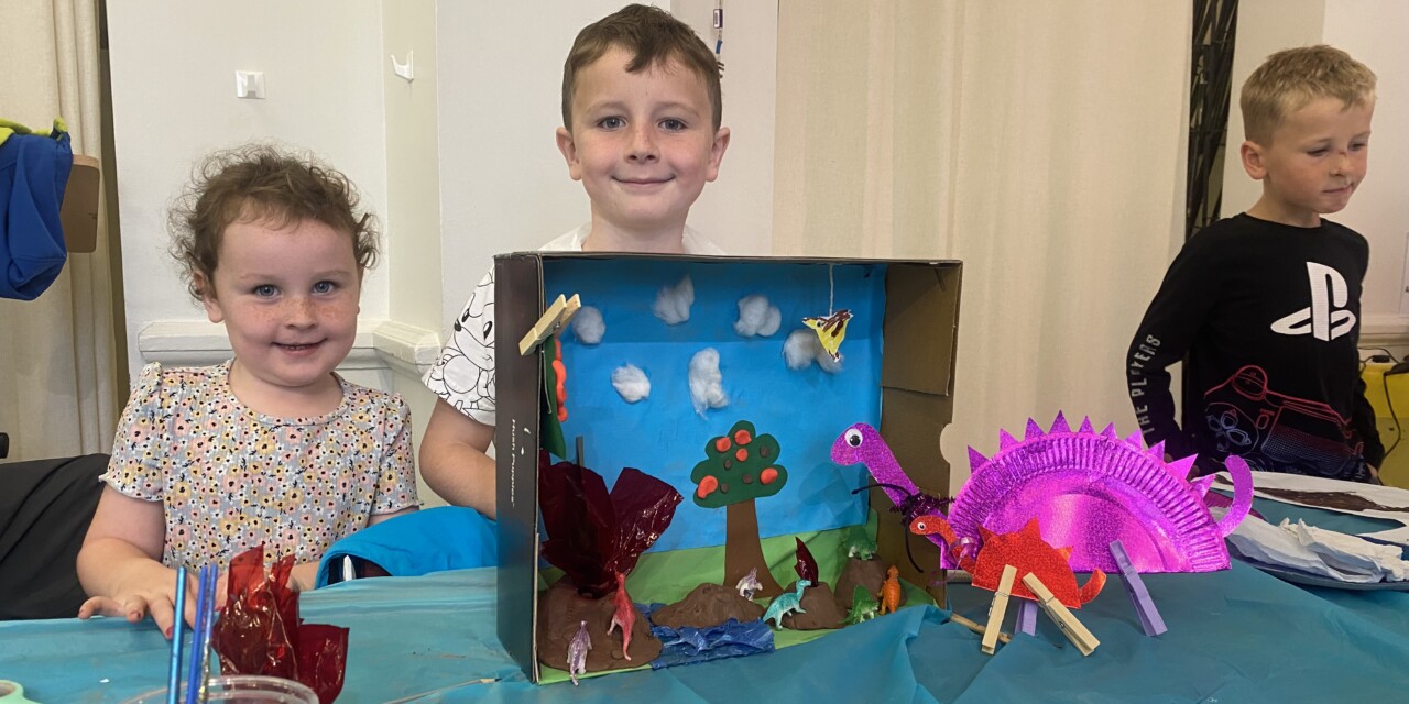 A photo of a boy and a girl standing proudly behind their dinosaur diorama, featuring two plate-o-saurus made from paper plates.