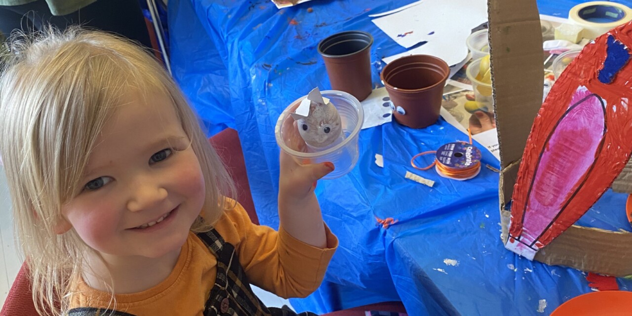 A young girl holds up an egg which she is decorating to look like an Easter bunny. There are a pair of bunny ears made from cardboard and painted on the table in front of her.