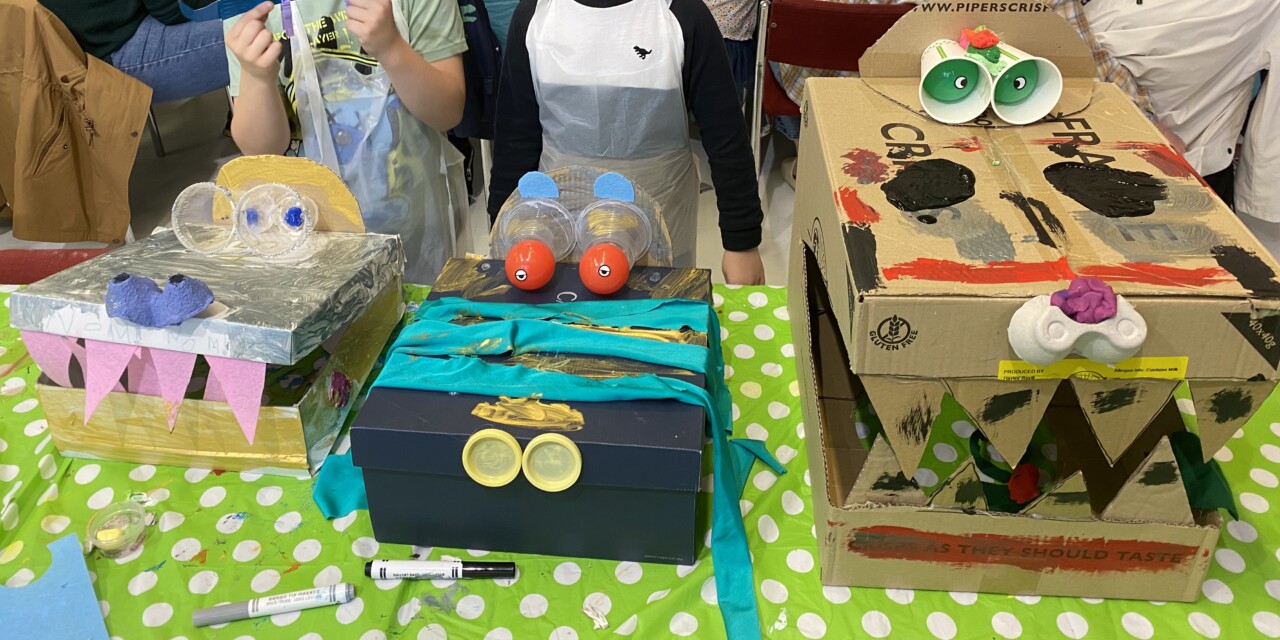 A photo of three dinosaur hats made out of cardboard boxes, paper cups and other recycled materials, decorated with paints, coloured pens and various crafting materials.