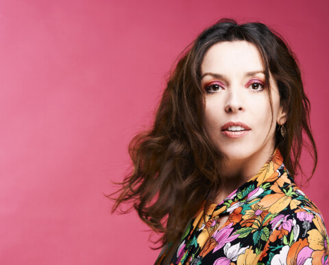 A photo of comedian Bridget Christie. She is standing in side profile but looking towards the camera. She is wearing a flower patterned dress, and set against a pink background.