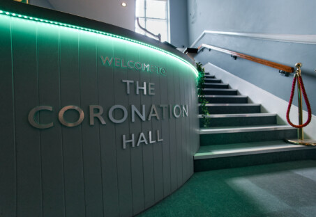 A photo of the reception desk at The Coro. It is made of vertical wooden slats painted in a grey-blue, with a dark wooden surface. 'Welcome to The Coronation Hall' is spelt out in silver lettering on the front and illuminated by a coloured downlight from above.