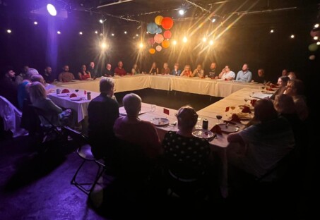 A photo of c.30 people sat around a set of tables laid out in a square with a space in the middle. They all have plates of food and drinks in front of them.